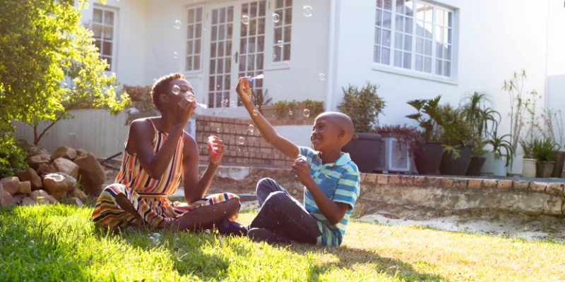 A mother and son sitting on green grass in a sunny backyard, blowing soap bubbles in front of their white residential Florida home.